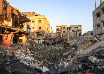 People stand on the edge of a crater caused by an Israeli bombardment as they inspect the destroyed building (L) of Palestinian journalist Adel Zorob, who was killed overnight, in Rafah in the southern Gaza Strip on December 19, 2023, amid continuing battles between Israel and the Palestinian Hamas militant group. Twenty Palestinians were killed in a bombing in Rafah on December 19, according to Hamas. Among them were four children and journalist Adel Zorob. (Photo by Mahmud HAMS / AFP)