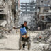 A boy pushes a wheelchair carrying water containers past the rubble of destroyed buildings along a street in Khan Yunis in the southern Gaza Strip on July 8, 2024 amid the ongoing conflict in the Palestinian territory between Israel and Hamas. (Photo by Bashar TALEB / AFP)