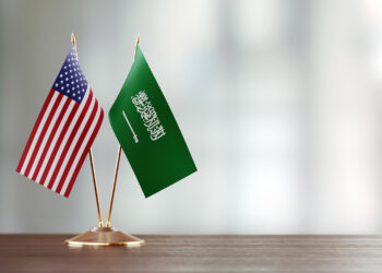 American and Saudi Arabian flag pair on desk over defocused background. Horizontal composition with copy space and selective focus.