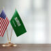 American and Saudi Arabian flag pair on desk over defocused background. Horizontal composition with copy space and selective focus.