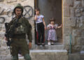 An Israeli soldier stands watch in the Old City of Hebron, West Bank, as two young children look on from the entrance of their home on September 14, 2024. (Photo by Mosab Shawer / Middle East Images / Middle East Images via AFP) (Photo by MOSAB SHAWER/Middle East Images/AFP via Getty Images)