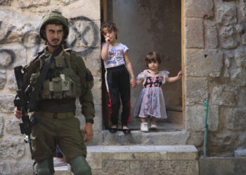 An Israeli soldier stands watch in the Old City of Hebron, West Bank, as two young children look on from the entrance of their home on September 14, 2024. (Photo by Mosab Shawer / Middle East Images / Middle East Images via AFP) (Photo by MOSAB SHAWER/Middle East Images/AFP via Getty Images)