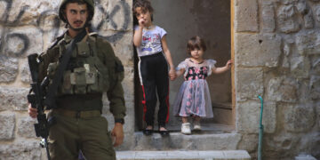 An Israeli soldier stands watch in the Old City of Hebron, West Bank, as two young children look on from the entrance of their home on September 14, 2024. (Photo by Mosab Shawer / Middle East Images / Middle East Images via AFP) (Photo by MOSAB SHAWER/Middle East Images/AFP via Getty Images)