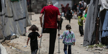 Abdullah Abu Harbeed walks on crutches with his daughters at a refugee camp in Gaza City, Gaza Strip, on June 27, 2026. Abdullah Abu Harbeed lost a leg in a 2014 strike and sustained severe injuries to his hand, abdomen, and remaining leg in a 2023 strike. He was later detained by the Israeli military, where he reports being subjected to beatings that dislodged metal fixation rods in his hand. Displaced from Beit Hanoun, he now lives in a tent with his family under difficult humanitarian conditions. (Photo by Abood Abusalama / Middle East Images / AFP via Getty Images)
